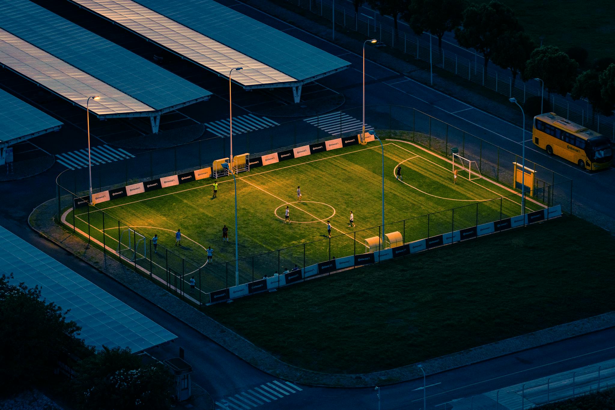 Aerial shot of a soccer field lit at night with players, surrounded by urban structures.